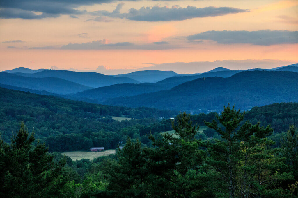 Sunset over Layered Blue Mountains with Forest in the Foreground and a Small Red Barn in a Meadow