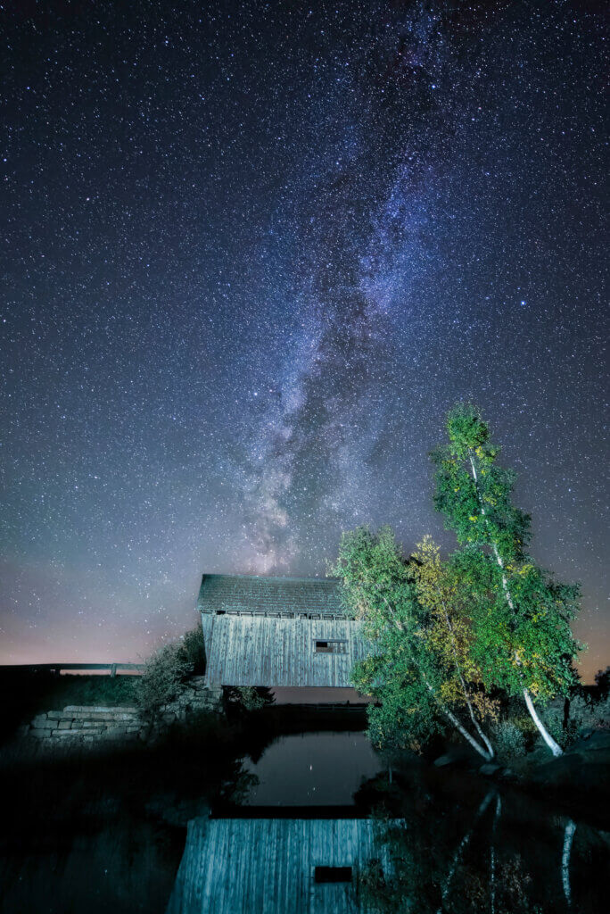 Milky Way Stretches Across a Starry Night over a Weathered Barn Beside a Calm Pond with Trees Reflecting in the Water
