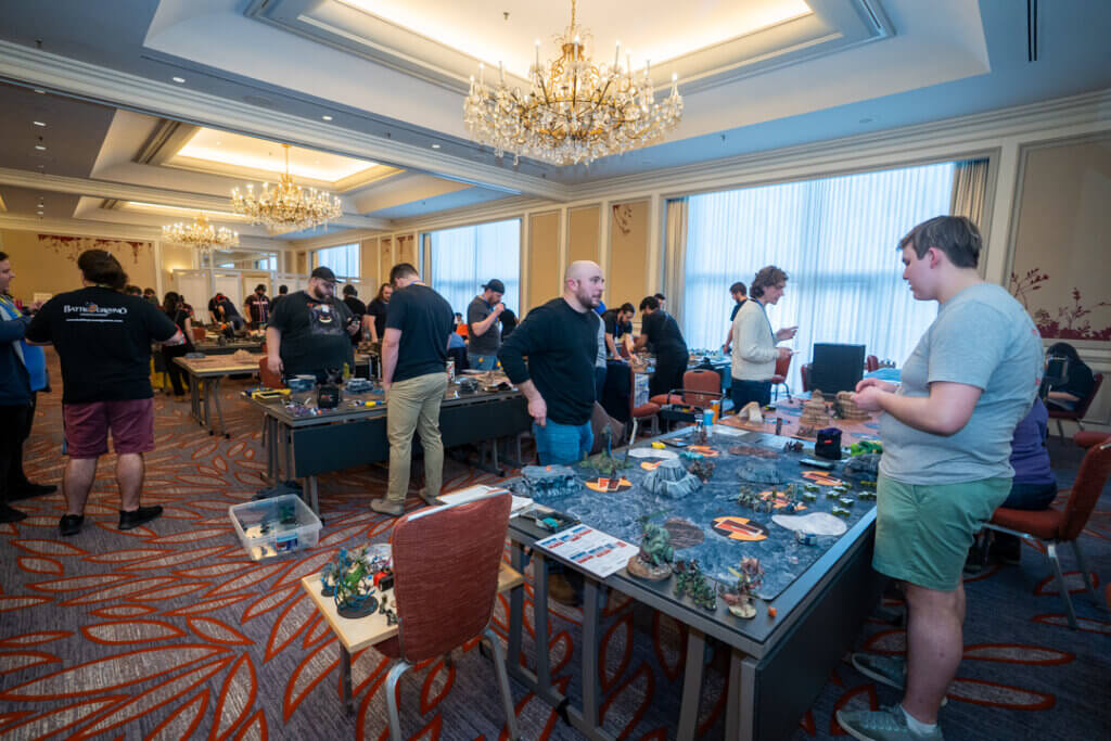 People Gathered Around Long Tables Playing Tabletop Miniature Wargames in a Hotel Conference Room with Chandeliers and Warm Lighting