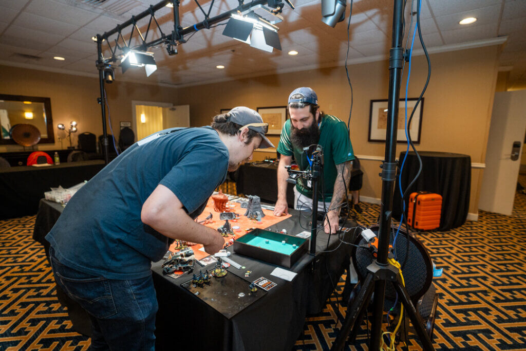 Dsc08735 Wicked Dicey Two Men in a Studio Set Up Around a Tabletop Game with Miniature Figures and a Green Dice Tray Under Bright Video Lights
