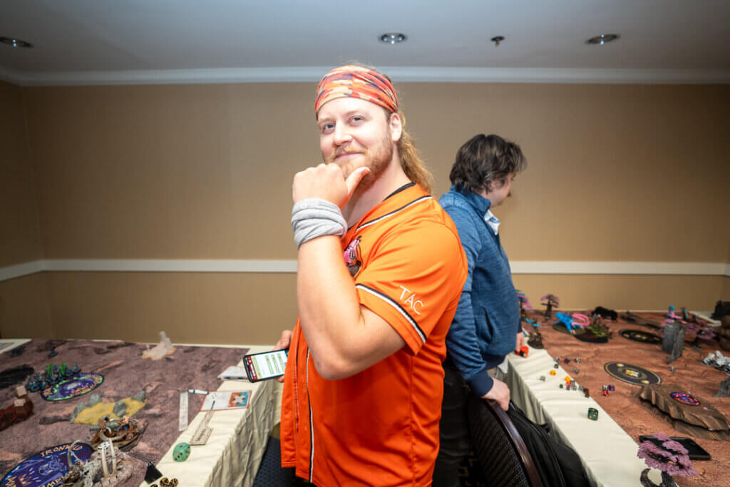 Age of Sigmar Tournament Wicked Dicey Smiling Man in an Orange Shirt and Striped Bandana Posing with His Hand on His Chin at a Tabletop Miniature Game Setup