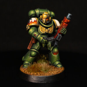 Green Space Marine miniature in ornate armor, holding a red weapon, with a skull emblem on the chest, standing on a grassy base.
