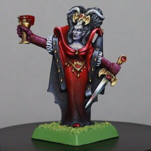 Painted tabletop miniature of a vampiric queen in a red gown with a black cape, horned crown, raised goblet in one hand and a dagger in the other, on a green base.