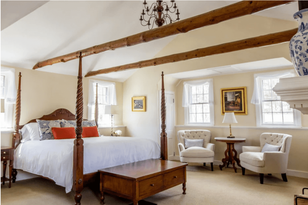 Hotel Bedroom with a Four poster Bed Exposed Wooden Beams and a Seating Area with Two Tufted Chairs and a Round Table Nearby