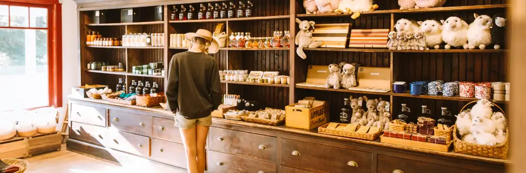 a Person in a Hat Browses Items Along Dark Wooden Shelves in a Cozy Shop Filled with Jars Mugs Chocolates and Plush Toys