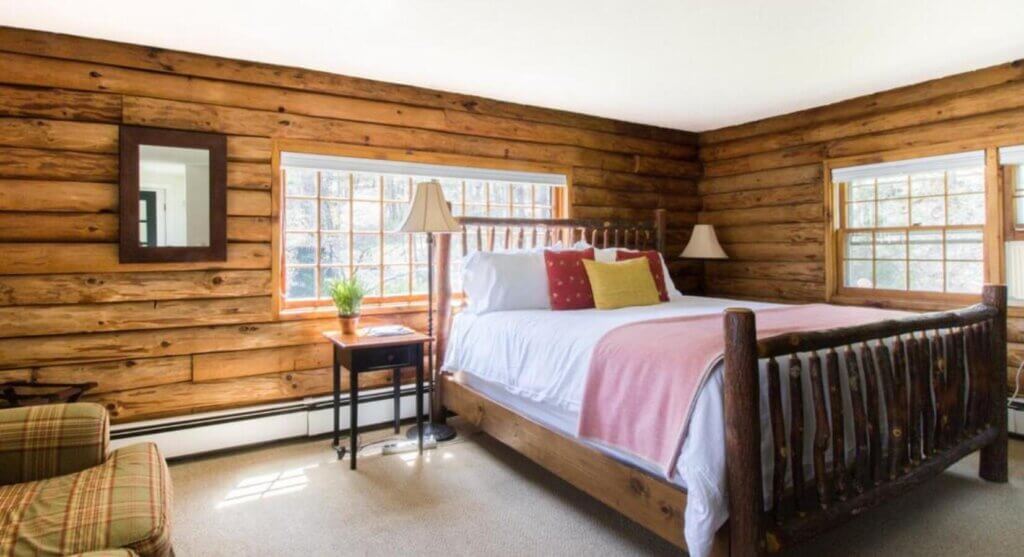 Rustic Wooden Bedroom with a Log Wall Queen Bed Pink Throw White Linens and Colorful Pillows by Two Nightstands and Lamps