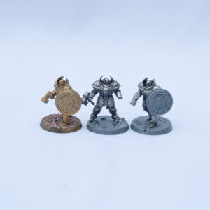 Three tabletop fantasy miniatures standing in a row: a bronze-painted warrior on the left with a round shield and hammer, flanked by two gray armored warriors with shields on bases.